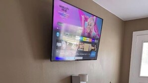 A small room with a television mounted on the wall displaying a program. There is a wooden bench, a small table with a coffee maker, and a refrigerator. - The Greenland Motel - Room No.3 (Munising)