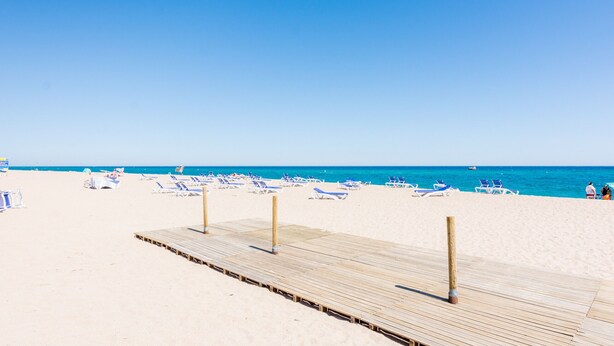 A wooden pier leading to a beach with blue chairs and a clear blue sky.