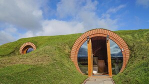 Una estructura circular de ladrillo con puerta de vidrio, situada sobre una colina cubierta de hierba bajo un cielo parcialmente nublado.