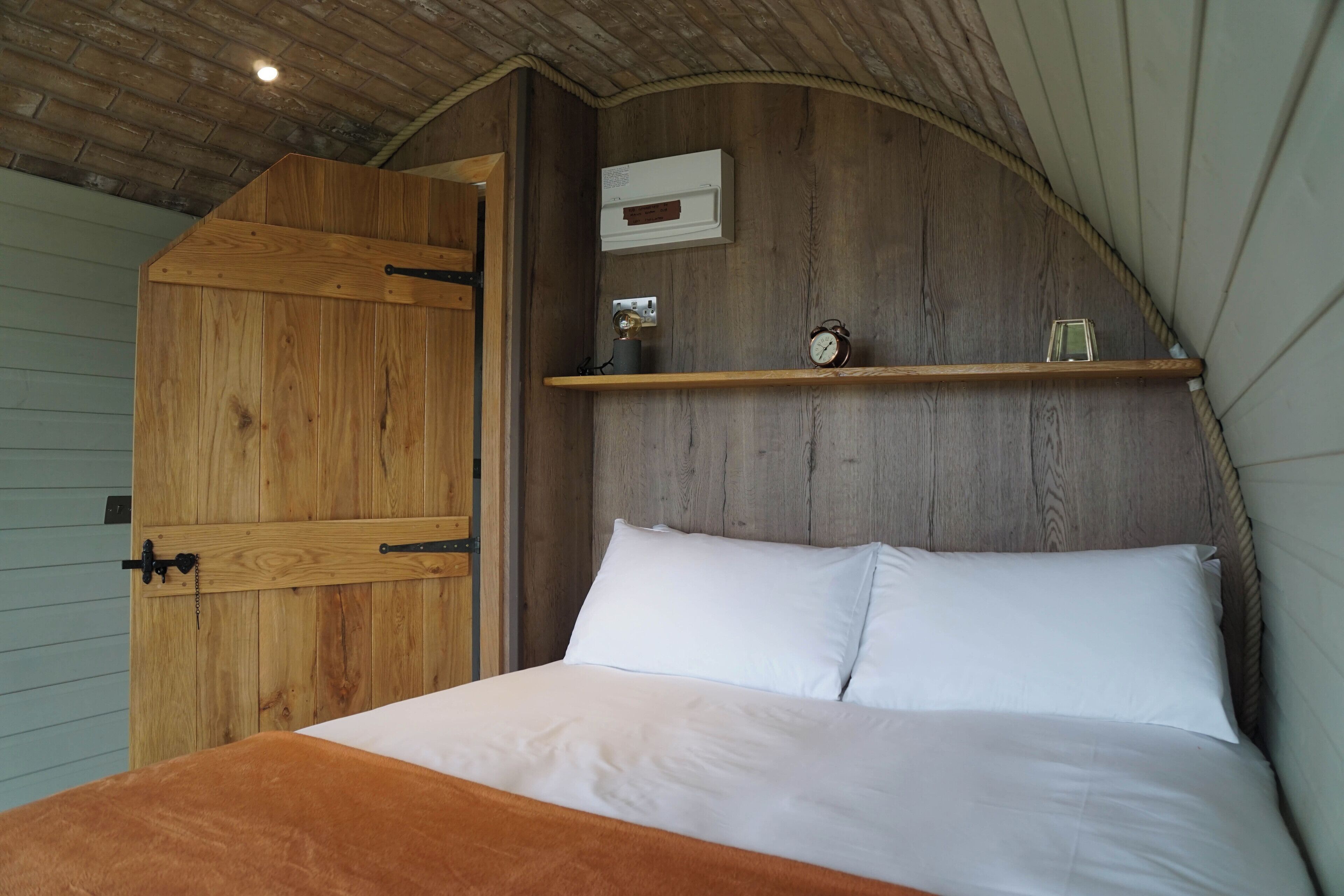 A bedroom with a wooden headboard, a bed with white linens, a shelf with a clock and a small decorative item, and a wooden door.