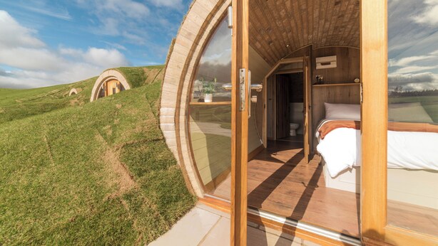Una estructura circular moderna de madera con una puerta de cristal que da a un dormitorio con cama y suelo de madera.