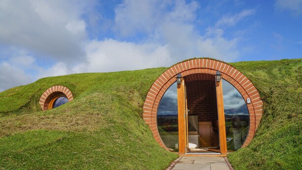 Una estructura circular de ladrillo con puerta de vidrio, situada sobre una colina cubierta de hierba bajo un cielo parcialmente nublado.