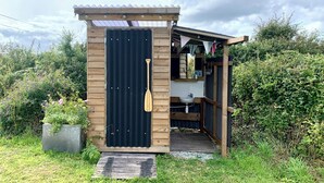 A small wooden structure with a black door, a wooden paddle hanging on the door, and a small sink inside. - Boutique Luxury Bell Tent Near Torpoint (Torpoint)