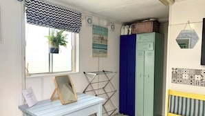 A small, well-lit room with a window, a blue and white patterned curtain, a mirror on a blue table, a drying rack, a blue wardrobe, a yellow bench, and a wall with a 'Beach Rules' sign. - Boutique Luxury Bell Tent Near Torpoint (Torpoint)