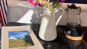 A neatly made bed with striped bedding, a white vase with pink flowers, a book titled "Welcome to Tregantle Farm Eco Glampsite," and a black lantern on a black table. - Boutique Luxury Bell Tent Near Torpoint (Torpoint)