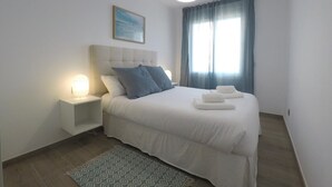A neatly arranged bedroom with a large bed, a bedside table, a wall-mounted lamp, and a window with curtains. - RECONNECTAR Malgrat de Mar Garbí I (Malgrat de Mar)