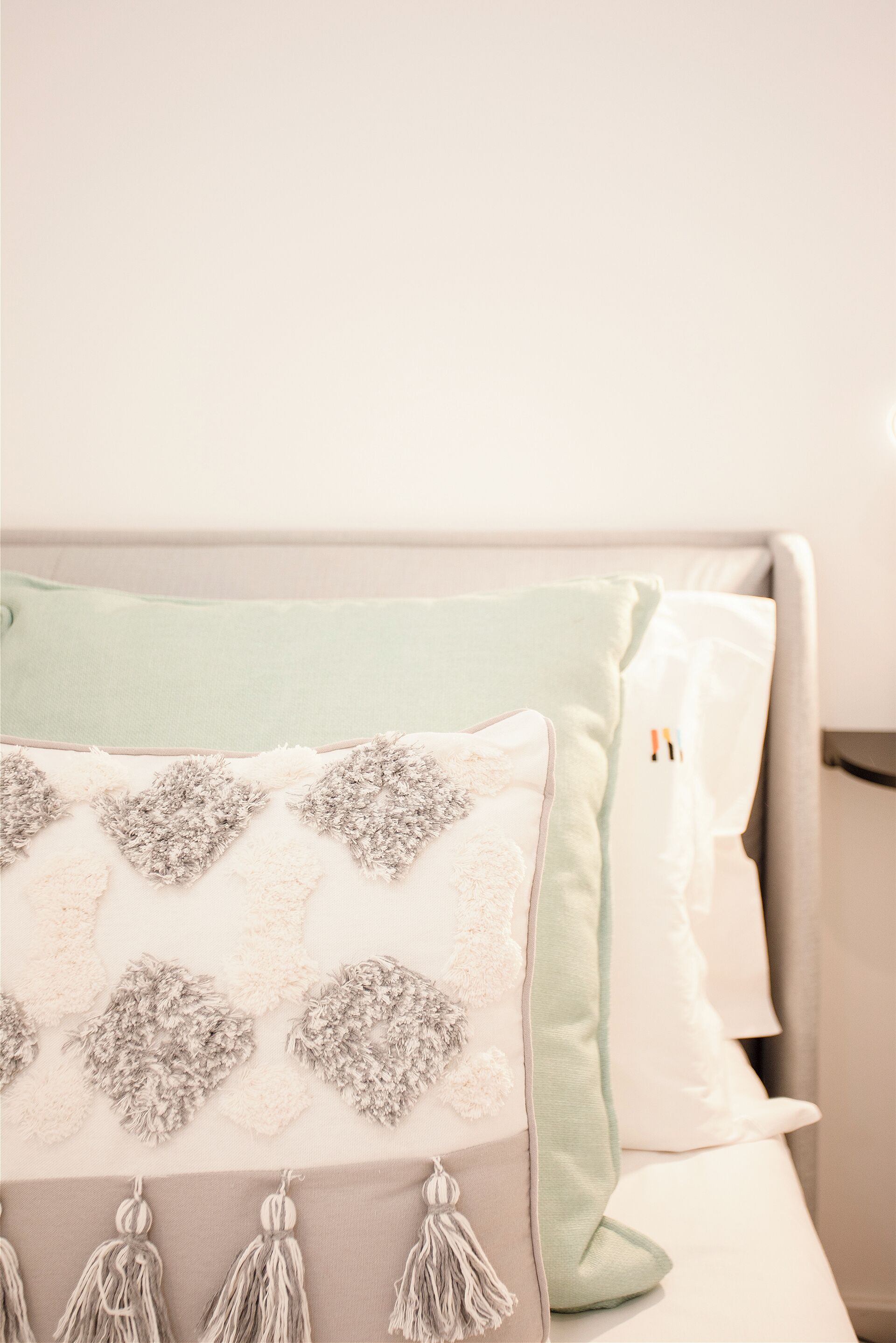 A bed with a light green throw blanket, a white pillow with a floral pattern, and a grey pillow with tassels.