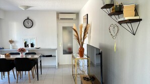 A modern living room with a TV, a dining area with chairs, a kitchen in the background, and a clock on the wall.