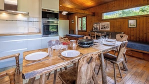 A wooden interior with a dining table set for four, a kitchen with modern appliances, and a large window offering a view of greenery. - Rustic chalet with garden in Chamonix (Chamonix-Mont-Blanc)