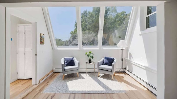 A room with a sloped ceiling, large windows, two armchairs, a small round table with a plant, and a white door. - Renovated Condo in Luxury Neighborhood (Stowe)