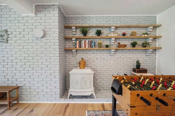 A room with a white brick wall, wooden shelves with books and plants, a white stove, and a foosball table.