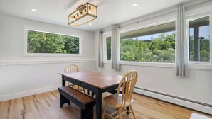 Un comedor con mesa y sillas de madera, ventanas amplias con vistas a zonas verdes, y una lámpara colgante moderna.