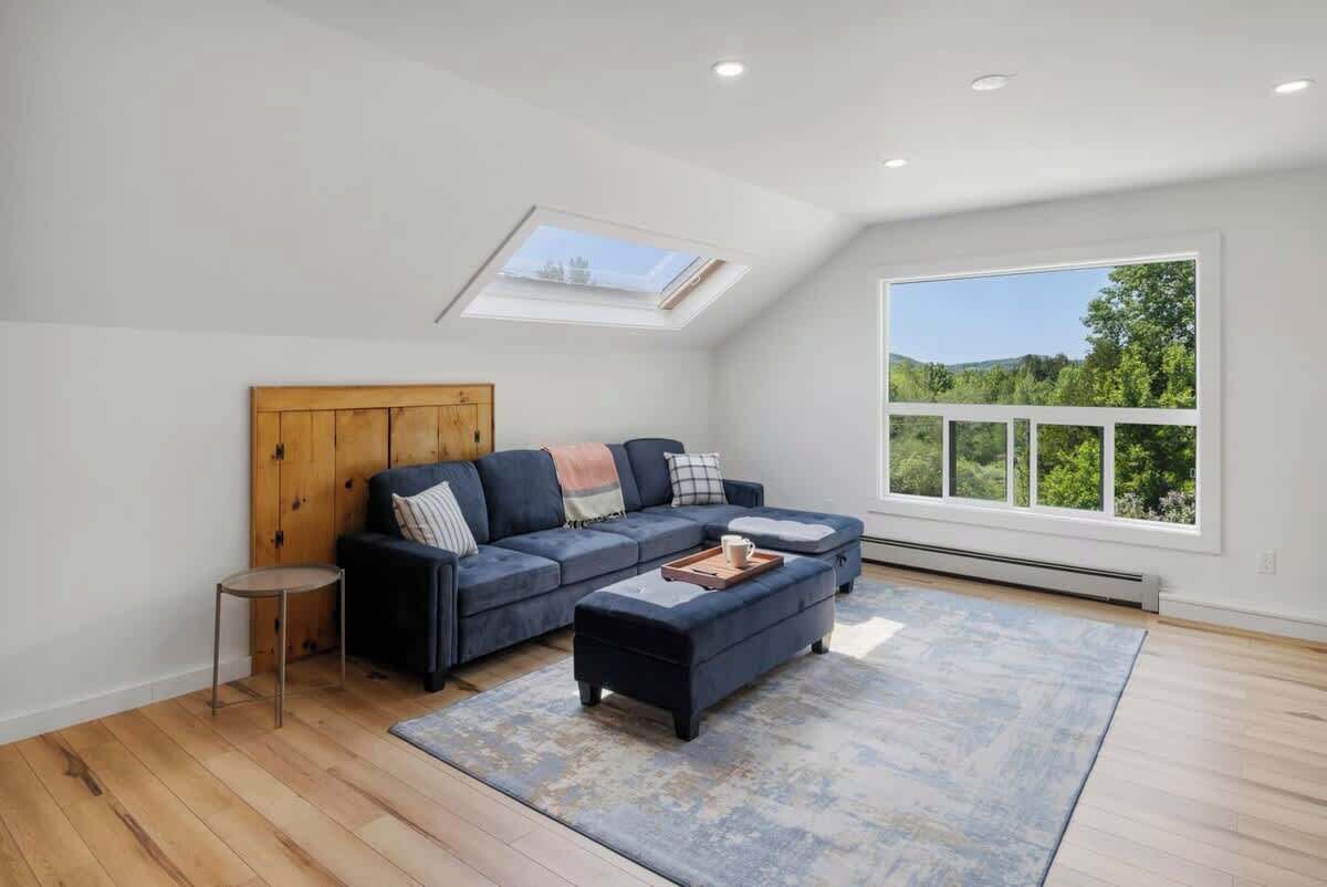 A spacious loft bedroom with a large skylight, a dark blue sectional sofa, a small round table, and a view of greenery through a large window.