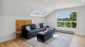 A spacious loft bedroom with a large skylight, a dark blue sectional sofa, a small round table, and a view of greenery through a large window.