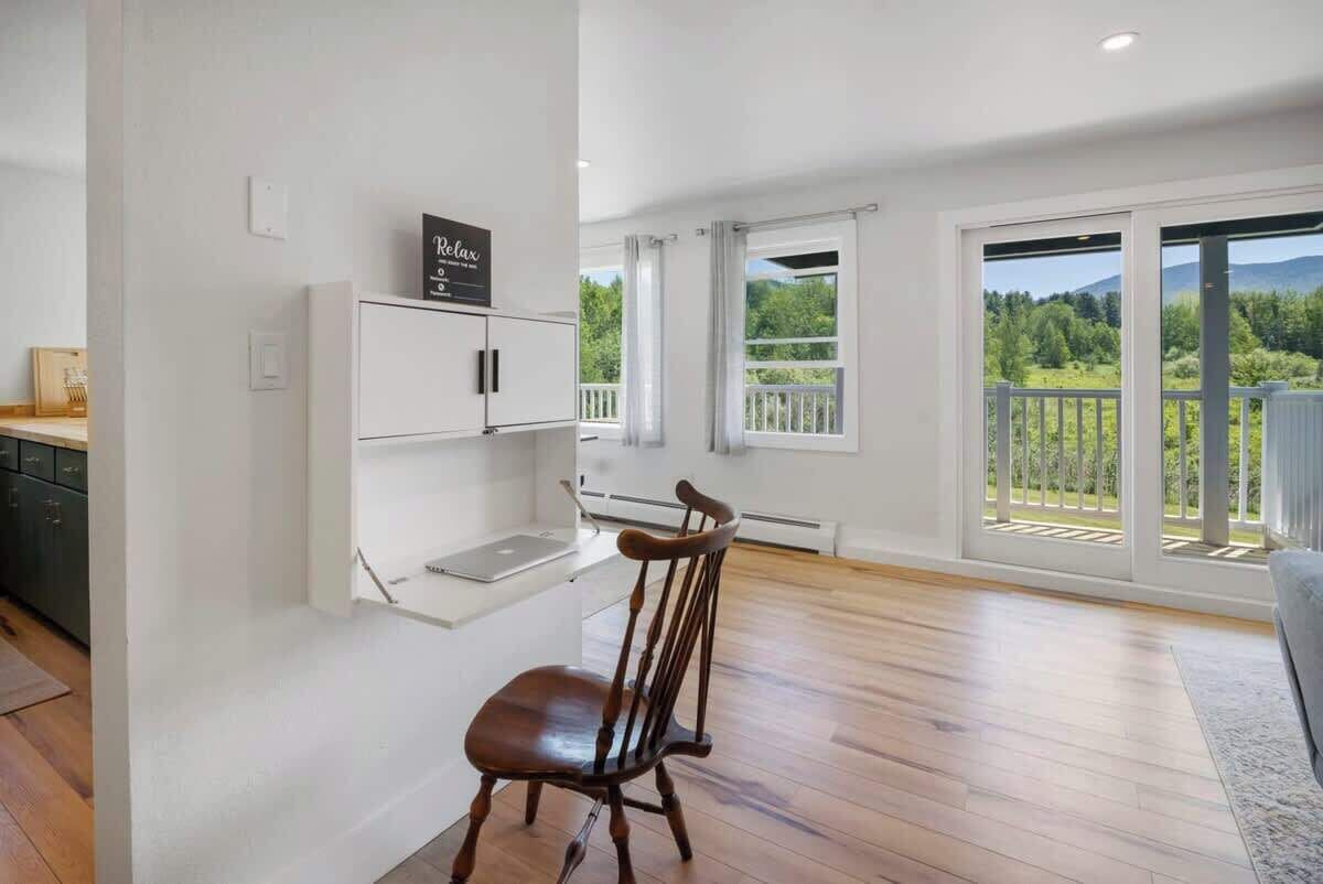 Una habitación amplia con piso de madera, un comedor con una silla, un estante blanco montado en la pared y vista a follaje a través de ventanas grandes.