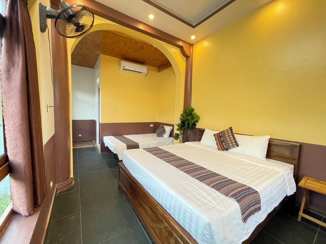 Een hotelkamer met twee bedden, een groot raam met gordijnen, een airconditioning en een kamerplant.