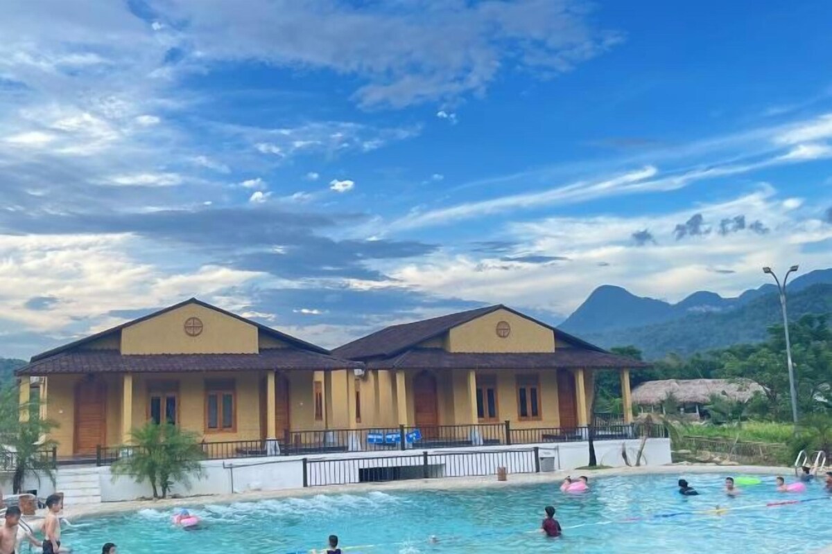 A swimming pool area with two yellow buildings, a mountain backdrop, and people swimming.