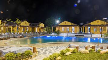 Un espace piscine nocturne agrémenté d’un chemin en pierre, de palmiers et de bâtiments aux toits jaunes.