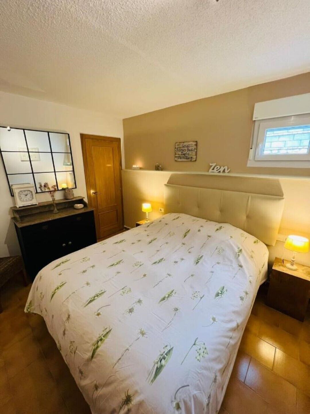 Ein Schlafzimmer mit einem großen Bett, einem Schminktisch, einem Spiegel, einer Nachttischlampe und einem Fenster mit Jalousien.