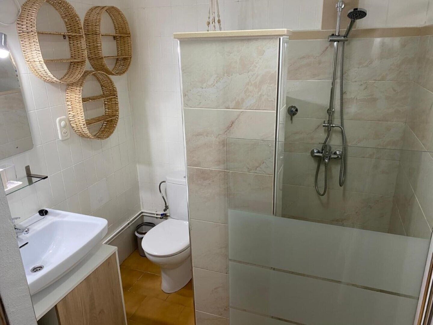 Un baño con ducha acristalada, un lavamanos blanco, un inodoro y dos adornos de mimbre en la pared.