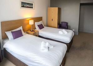 Two neatly made beds with white linens and purple pillows, a wooden wardrobe, a purple chair, and a framed picture on the wall. - Tor Park Hotel (Torquay)