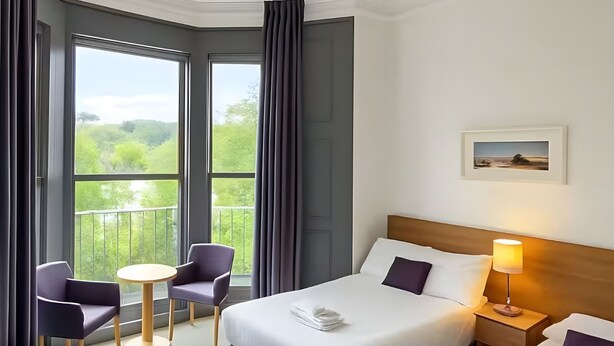 A hotel room with a large bed, a bedside table with a lamp, a chair, a small round table, and a view of greenery through large windows. - Tor Park Hotel (Torquay)