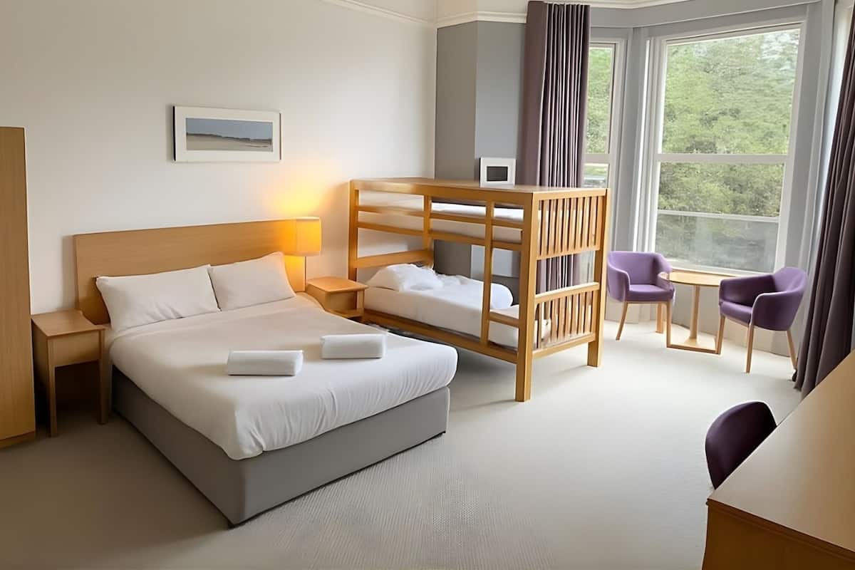 Hotellihuone, jossa on suuri sänky, puinen kerrossänky, työpöytä kahdella tuolilla ja ikkuna, jossa on verhot.