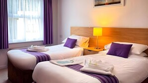 A hotel room with two beds, each with white linens and purple accents, a bedside table with a lamp, and a painting on the wall. - Tor Park Hotel (Torquay)