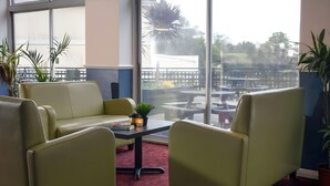 A room with a red carpet, two green sofas, a small table with a plant, and large windows offering a view of outdoor tables and chairs. - Tor Park Hotel (Torquay)