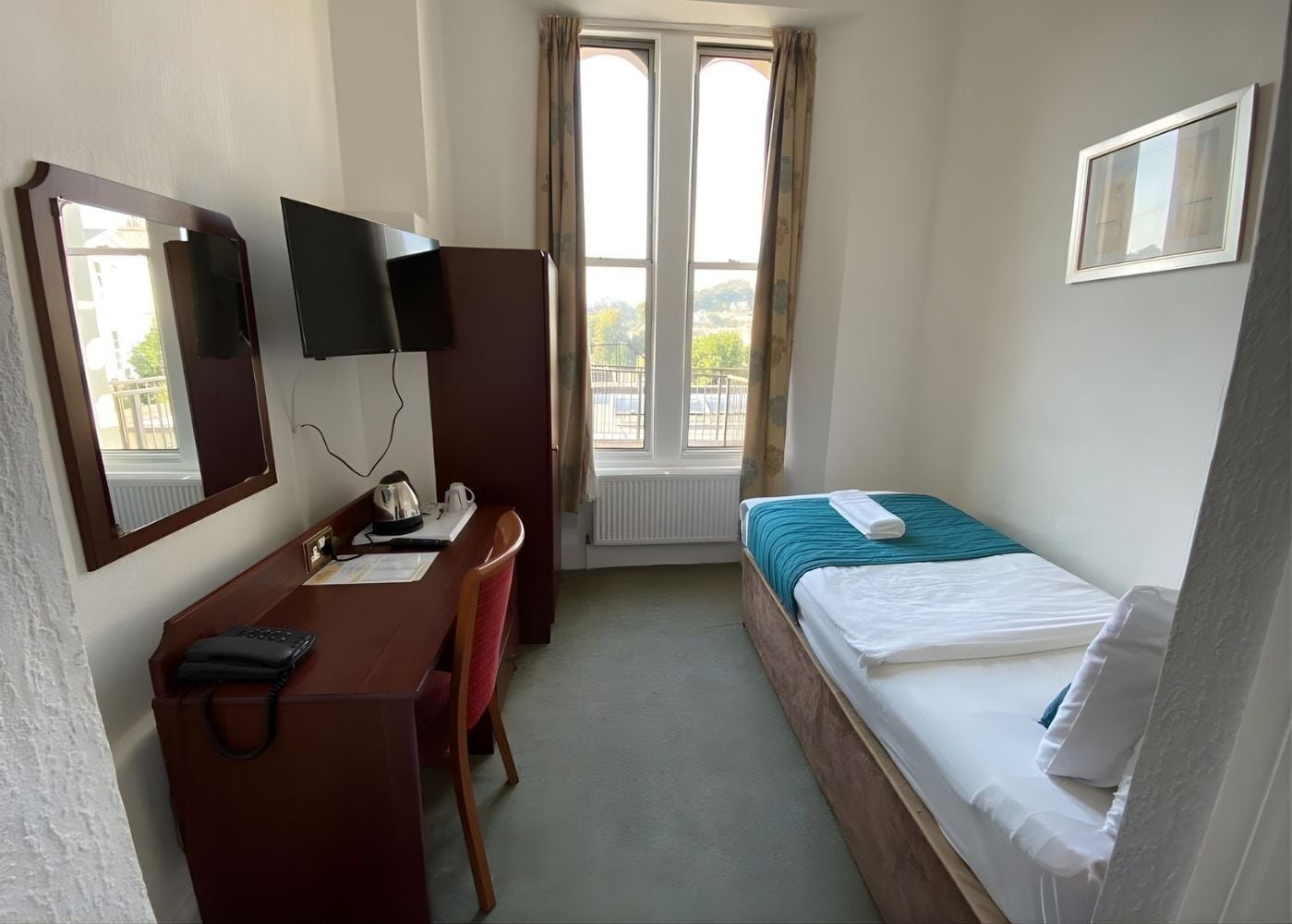 Ett litet hotellrum med en säng, ett skrivbord, en stol, en tv och ett fönster med gardiner.