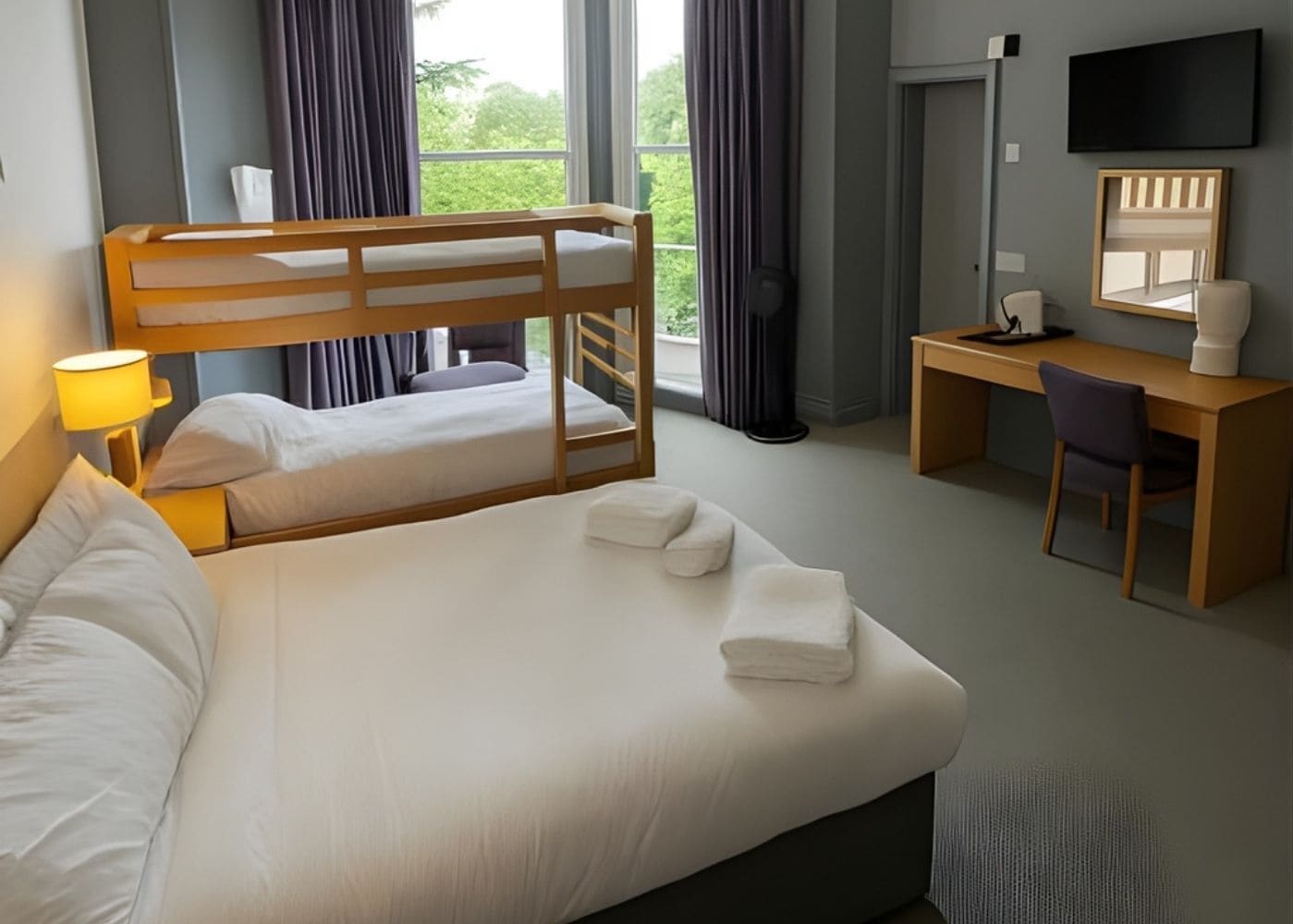 A hotel room with a bunk bed, a desk, a chair, a lamp, a TV, and a window with curtains.