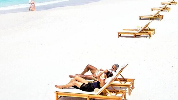 A beach with white sand and clear blue water. There are wooden lounge chairs and a couple walking on the beach. - Micro-Studio APT 5B, Oceanview + Pool, Near Beach | @ Paradise Point Barbados (Maxwell)