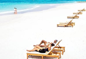 A beach with white sand and clear blue water. There are wooden lounge chairs and a couple walking on the beach.