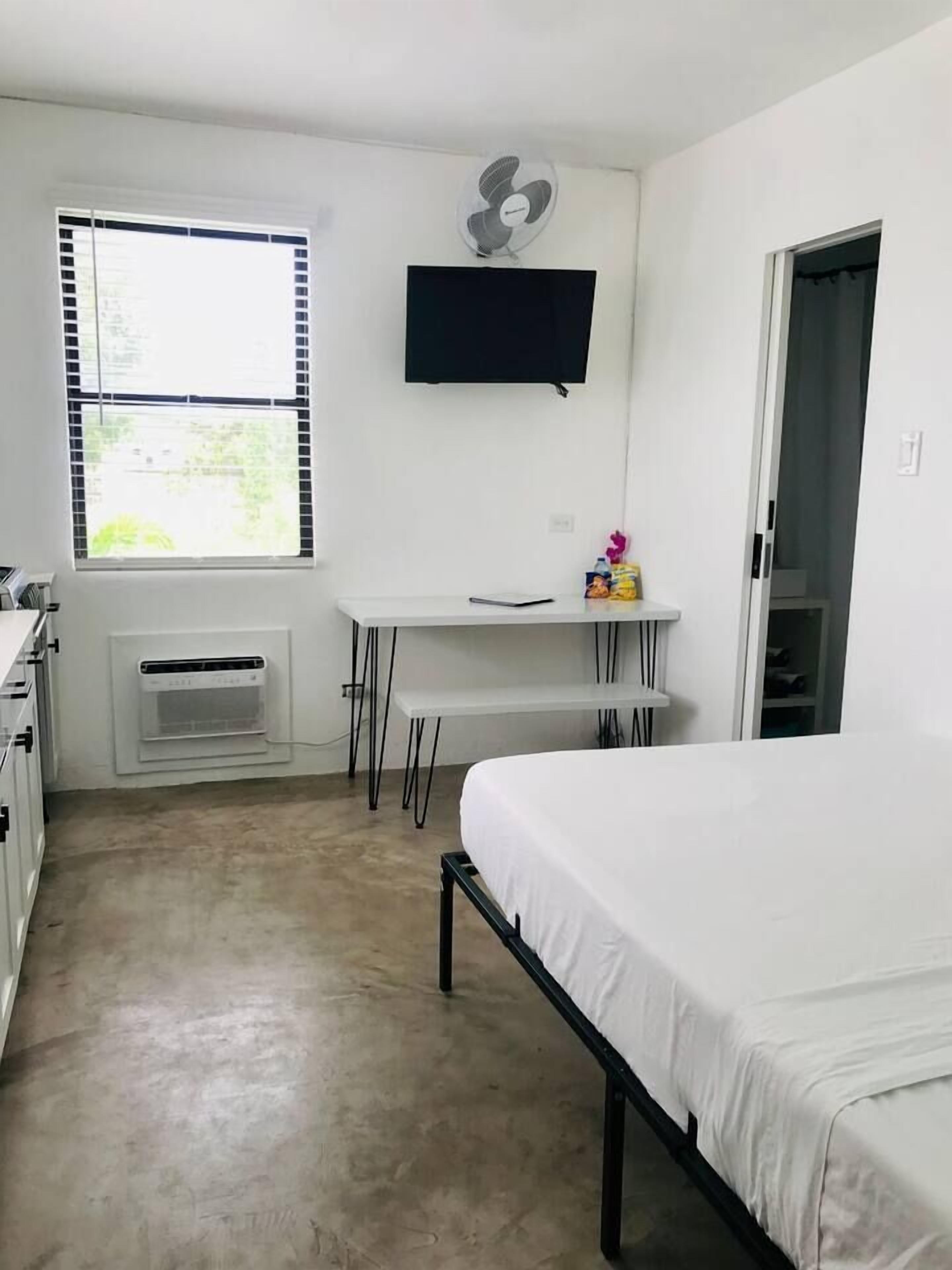 Ein Schlafzimmer mit einem Bett, einem Schreibtisch, einem Stuhl, einem Standventilator und einem Fenster mit Jalousien.