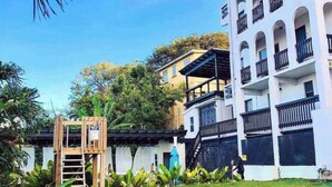 A multi-story building with balconies, a wooden play structure, and lush greenery. - Micro-Studio APT 5B, Oceanview + Pool, Near Beach | @ Paradise Point Barbados (Maxwell)