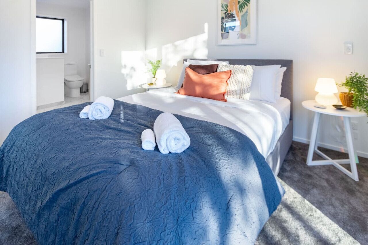 A neatly made bed with a blue textured bedspread, white pillows, and a single orange pillow. There are two rolled towels on the bed. A small white nightstand with a lamp and a plant is to the right of the bed.