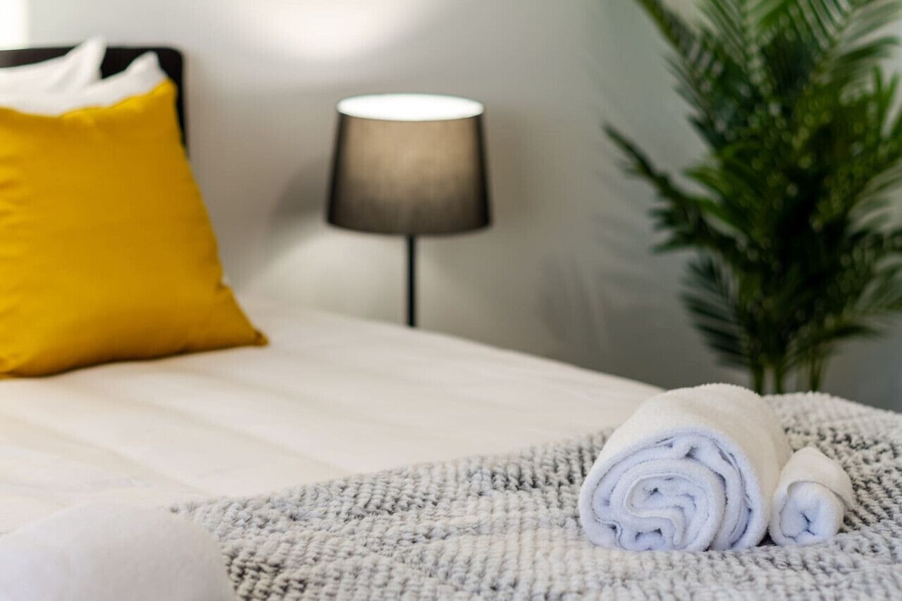 A neatly made bed with a rolled white towel, a yellow pillow, and a lamp in the background.