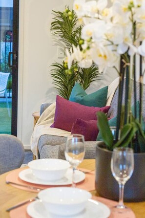 Zona pranzo con tavolo apparecchiato per due, un vaso di fiori bianchi e vista su un giardino attraverso una porta di vetro.