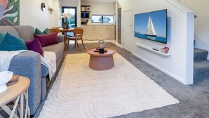 Een moderne woonkamer met een bank, salontafel en televisie. Achtergrond is een keuken met een raam.
