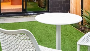 Een terras met een ronde tafel en twee witte stoelen, een houten schutting en een glazen deur die leidt naar een binnenruimte met een keuken en eethoek.