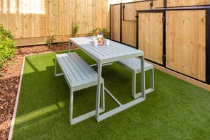 Een witte picknicktafel met bankjes in een achtertuin met kunstgras en een houten omheining.
