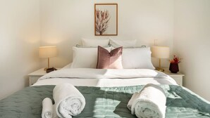 Een net opgemaakt bed met een groene sprei, twee opgerolde handdoeken en een ingelijst botanisch kunstwerk aan de muur.