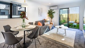 Een moderne woonkamer met een eethoek, een grijze bank, een salontafel en een groot raam met uitzicht op de natuur.