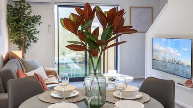 Een eetruimte met een ronde tafel voor vier personen, omringd door moderne stoelen, een vaas met rode bladeren, en een televisie waarop een stadsbeeld te zien is.
