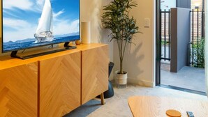 Una sala moderna con un aparador de madera, un televisor mostrando un velero, una planta en maceta y vistas al exterior.