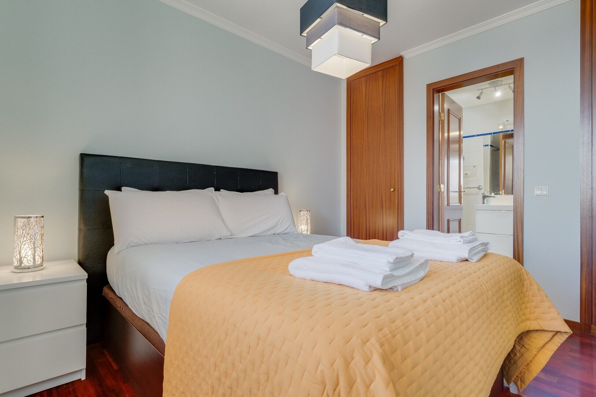 A neatly made bed with a beige quilt, a wooden headboard, and white bedding. There is a bedside table with a lamp and a mirror on the wall.