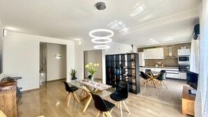 A modern living room with a glass dining table, black chairs, and a yellow sofa. There is a kitchen in the background with white cabinets and a black countertop.