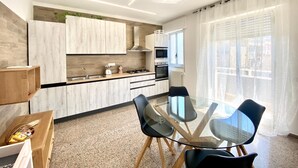 A modern kitchen with white cabinets, a gas stove, and a microwave. There is a dining table with chairs and a window with sheer curtains.