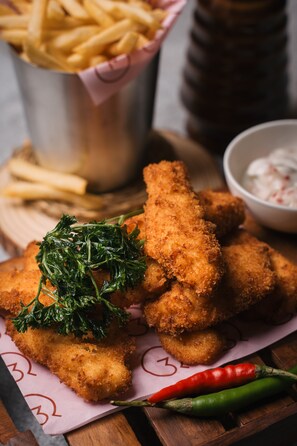 Un plato de comida rebozada y frita, posiblemente pollo, adornado con hierbas, acompañado de un recipiente con salsa para mojar y una porción de papas fritas.