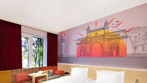 Ein Hotelzimmer mit einem großen Bett, einem Sofa, einem kleinen Tisch und einem Nachttisch. Ein Fenster mit Vorhängen und ein Wandbild eines Gebäudes sind ebenfalls vorhanden.